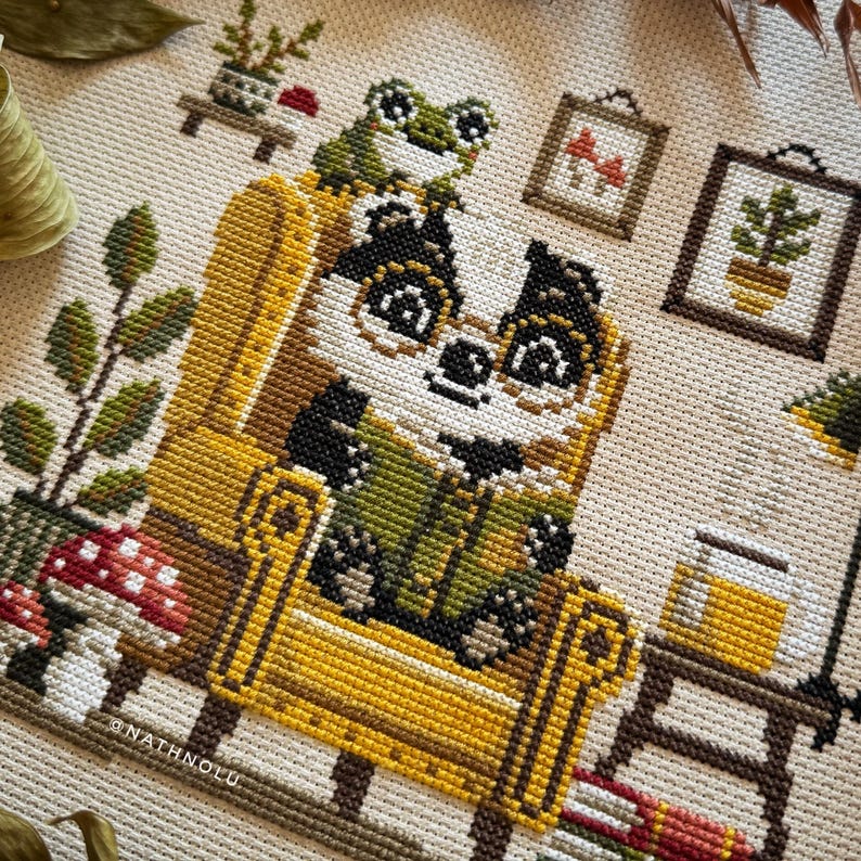 Cute badger cross stitch design sitting in a yellow chair surrounded by books, mushrooms, and cozy decor.