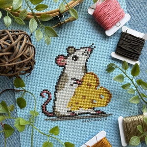 May include: Cross-stitch embroidery of a white mouse holding a wedge of yellow cheese. The background is light blue and the mouse is surrounded by green foliage.