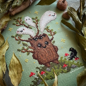 May include: Cross stitch pattern featuring a brown tree stump with a smiling face and three white ghosts with happy faces. The tree stump is surrounded by green leaves and red mushrooms. A black bird with a red beak is perched on a mushroom to the right of the tree stump.