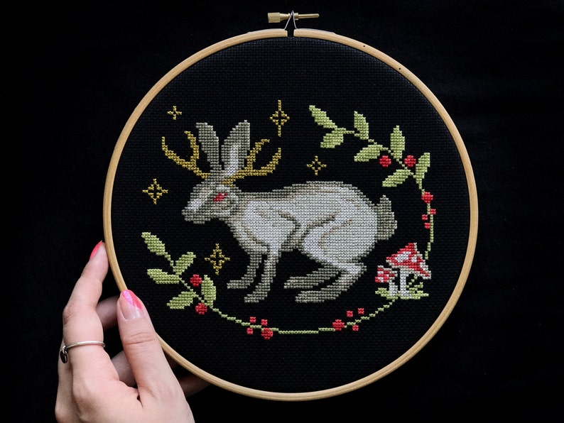 Cross stitch mythical Easter Bunny with horns on a black canvas in an embroidery frame that holds a hand on a dark background