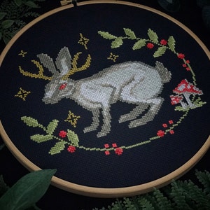Cross stitch mystical Easter bunny on black canvas. The hare has red eyes and shimmering horns. Photo taken in dim light. Around the embroidery plants on a dark background