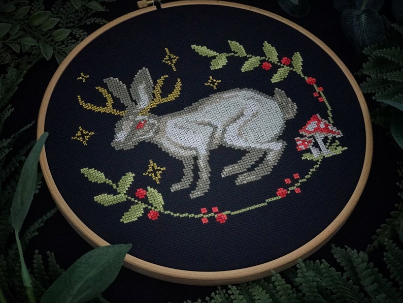 Cross stitch mystical Easter bunny on black canvas. The hare has red eyes and shimmering horns. Photo taken in dim light. Around the embroidery plants on a dark background