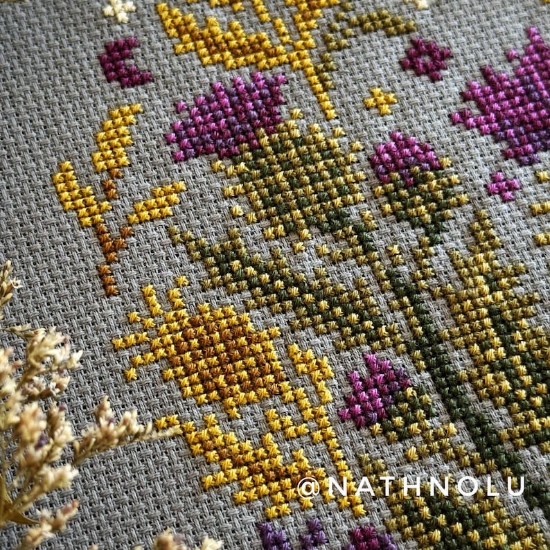 Detailed flower cross stitch showing purple thistle blooms and a realistic bee in a balanced ornamental layout.