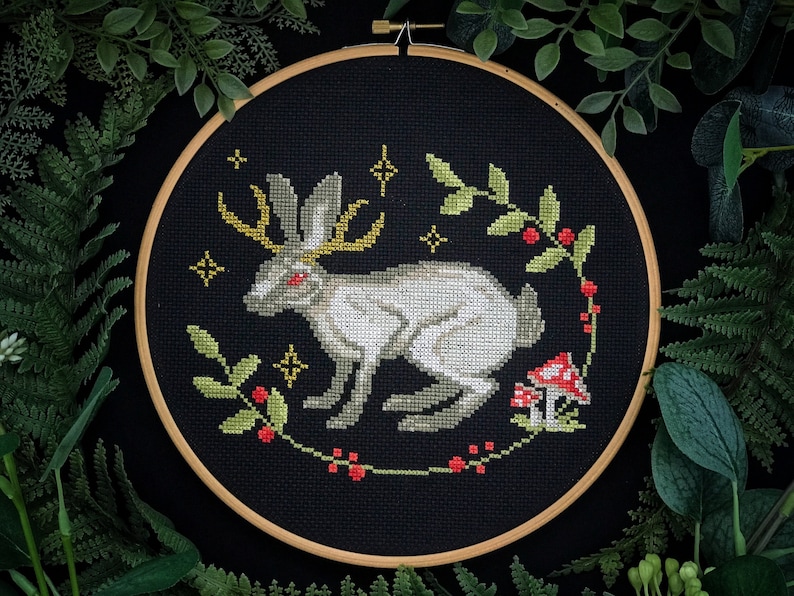 May include: A white rabbit with antlers and red eyes is embroidered on a black fabric background. The rabbit is surrounded by a wreath of green leaves and red berries. The background is black with a subtle texture.