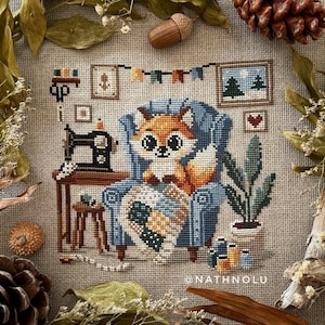 May include: A cross-stitch design featuring a cozy scene with a fox in a blue armchair, holding a blanket. The room includes a sewing machine, framed art, and a potted plant. The color palette is warm, with browns, blues, and oranges.