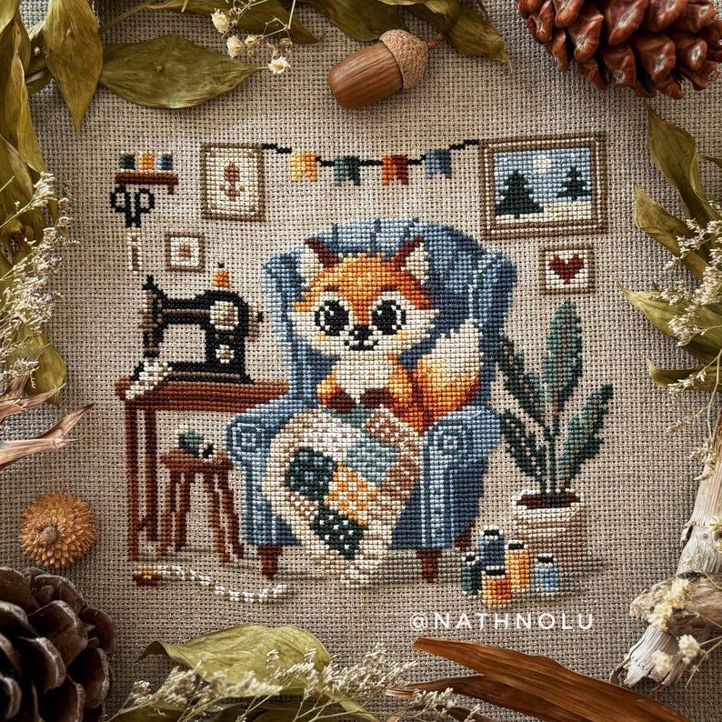Cross stitch pattern of a cute fox sitting in a large blue armchair while quilting a colorful patchwork blanket.
