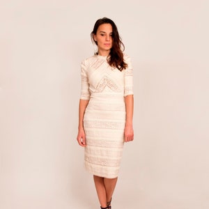 May include: A cream-coloured, knee-length dress with intricate lace detailing. The dress features a high neckline, three-quarter sleeves, and a fitted silhouette. The model is wearing black strappy heels. The background is a plain, light-coloured wall.