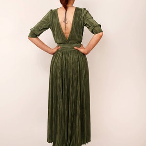 May include: Olive green pleated maxi dress with a plunging back and three-quarter sleeves. The dress features a fitted waist and a flowing skirt. The model is wearing black high-heeled boots. The dress is made of a shiny, textured fabric.