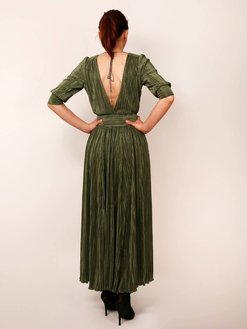 May include: Olive green pleated maxi dress with a plunging back and three-quarter sleeves. The dress features a fitted waist and a flowing skirt. The model is wearing black high-heeled boots. The dress is made of a shiny, textured fabric.