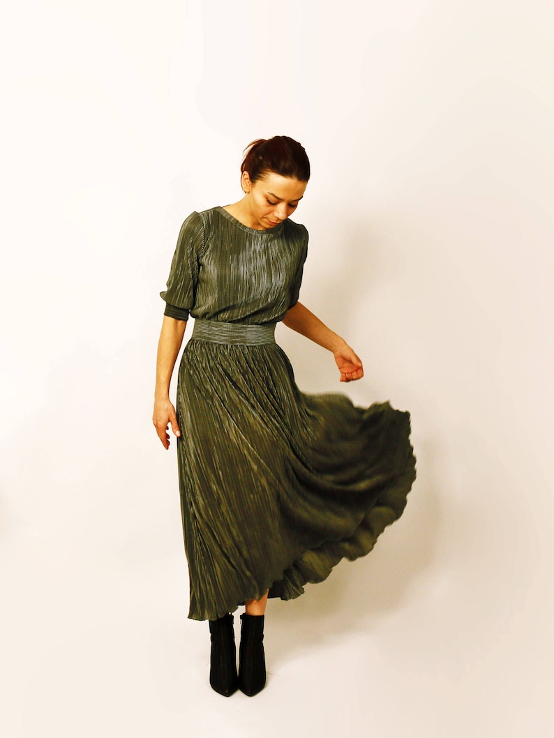 May include: An olive green pleated dress with short sleeves and a defined waist. The dress flows down to a midi length, paired with black ankle boots. The model is standing in front of a white background.