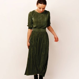 May include: Olive green pleated dress with short sleeves and a fitted waist. The dress has a round neckline and a flowing skirt. The model is wearing black ankle boots. The dress is made of a shiny, textured fabric.