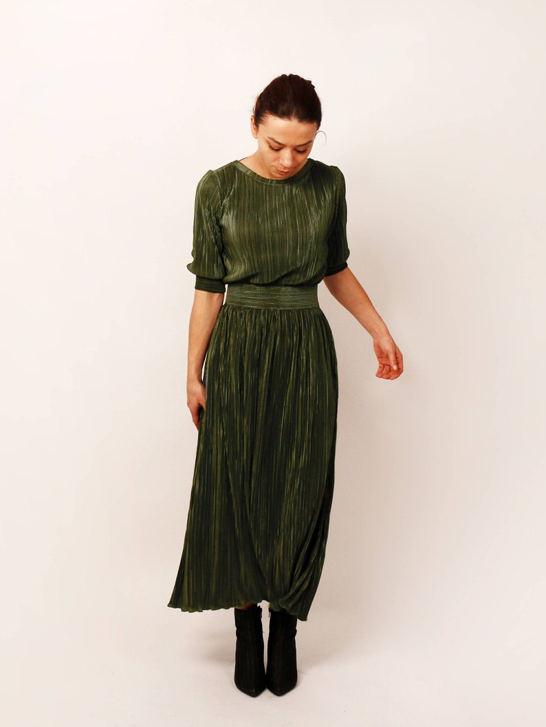 May include: Olive green pleated dress with short sleeves and a fitted waist. The dress has a round neckline and a flowing skirt. The model is wearing black ankle boots. The dress is made of a shiny, textured fabric.