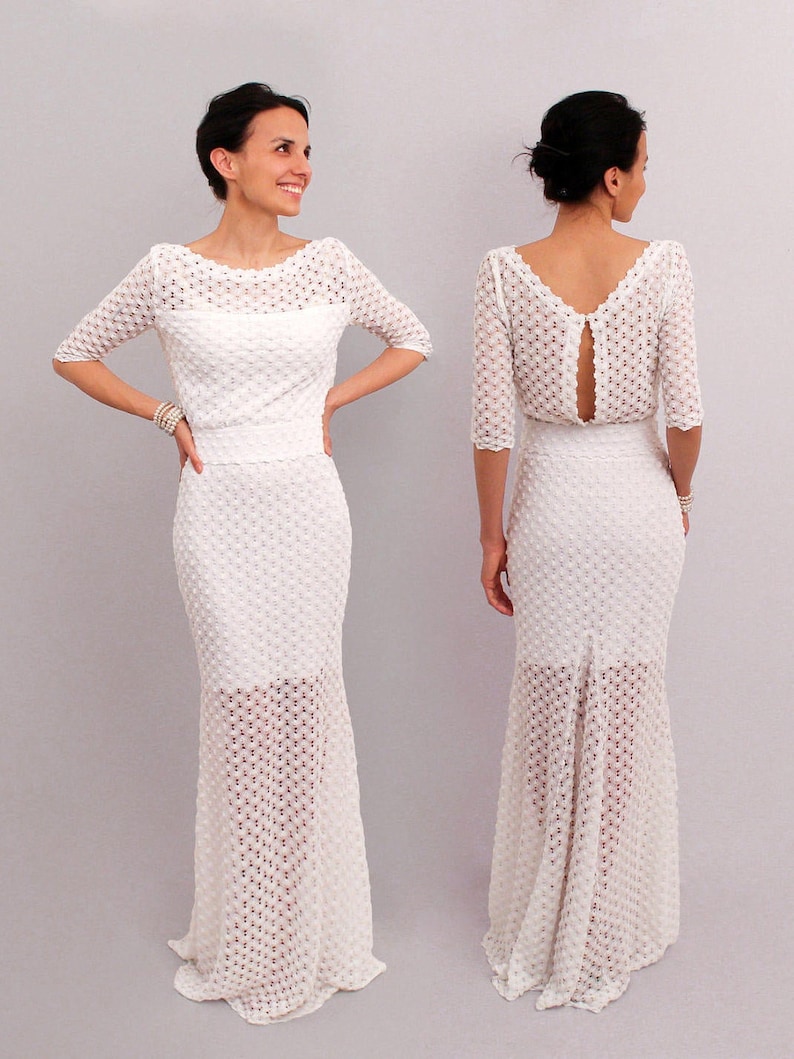 May include: A white, floor-length dress with a textured, open-weave design. The dress features a fitted bodice with short sleeves and a boat neckline. The skirt flares out at the bottom, creating a mermaid silhouette. The back of the dress has a keyhole opening.