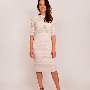 May include: A cream lace dress with a high neckline and three-quarter sleeves. The dress has detailed lace patterns and a knee-length skirt. The model is wearing black strappy heels.