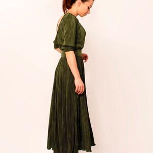 May include: Olive green pleated dress with a cinched waist and elbow-length sleeves. The dress has a flowing skirt and is paired with black ankle boots. The fabric has a subtle sheen, and the dress is designed with a low back.