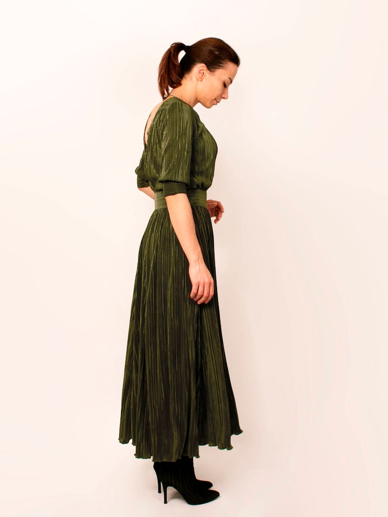 May include: Olive green pleated dress with a cinched waist and elbow-length sleeves. The dress has a flowing skirt and is paired with black ankle boots. The fabric has a subtle sheen, and the dress is designed with a low back.