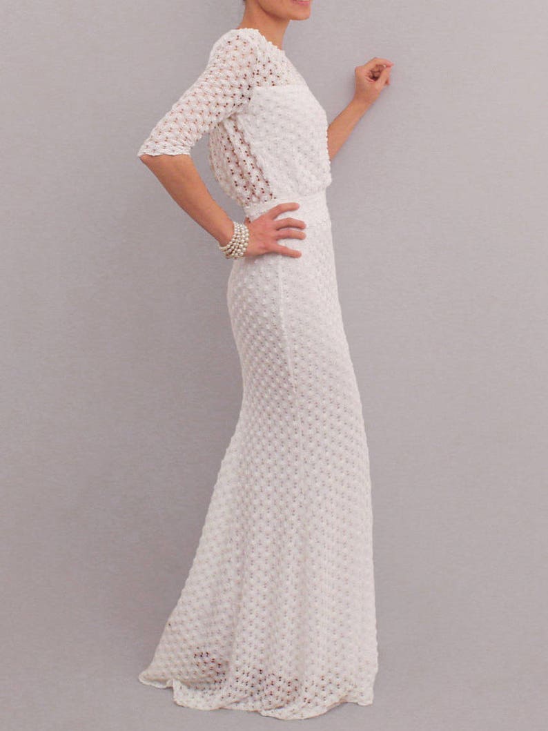 May include: A white, textured, floor-length dress with a matching bolero jacket. The dress features a fitted bodice and a flared skirt. The bolero has three-quarter sleeves. A pearl bracelet adorns the wrist. The dress is made of a delicate, open-weave fabric.