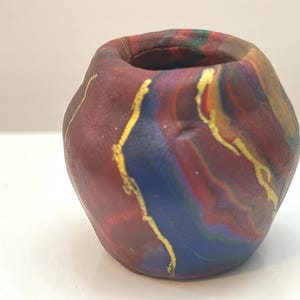 May include: A small, handmade ceramic vase with a unique, organic shape. The vase features a blend of colors, including deep red, blue, and hints of green and yellow, with gold accents. The opening is circular, and the vase is approximately 3 inches tall.