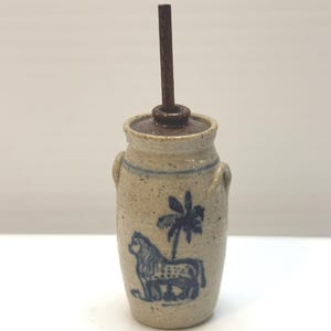 May include: A small, antique-style stoneware butter churn with a dark brown handle. The churn is beige with blue painted details, including a lion and palm tree design. It has a lid and two small handles.