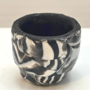 May include: A small, handmade ceramic cup with a unique zebra stripe pattern in black and white. The interior is a deep blue, and the rim is dark. The cup has a textured, organic shape, perfect for holding small items or as a decorative piece.