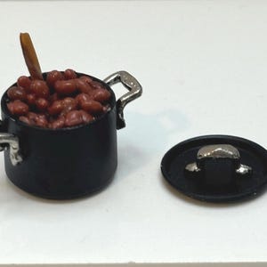 May include: Miniature black pot filled with red beans and a wooden stick, with a detached black lid. The pot has silver handles. This miniature food item is likely for a dollhouse or other small-scale display.