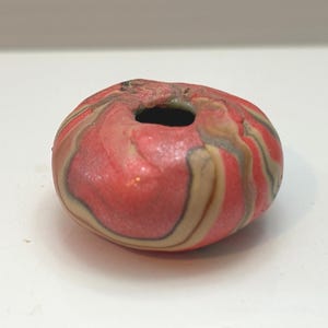 May include: A small, round, decorative object with a hole in the center. The object is primarily red with swirling patterns of tan and gray. The surface appears slightly textured and glossy. The overall shape is reminiscent of a small, flattened sphere.