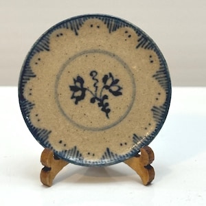 May include: A small, round, tan-colored plate with a blue floral design and scalloped edge. The plate is supported by a small wooden stand. The plate's rim has a blue, scalloped design. The center of the plate features a blue floral motif.
