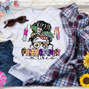 May include: A white t-shirt featuring a graphic of a person with a messy bun, wearing glasses with one eyeball and one patterned lens. The design includes a syringe, a test tube, and the text PHLEBOTOMIST LIFE in colorful, patterned letters.