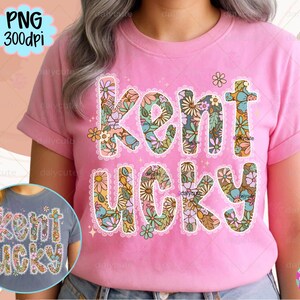 May include: A pink t-shirt with the words "Kent Ucky" in a floral design. The letters are filled with colorful flowers and outlined with a white lace-like trim. The shirt has short sleeves.
