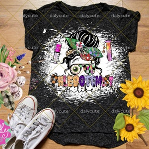 May include: A dark gray heathered t-shirt featuring a graphic of a messy bun hairstyle with a patterned headband, a syringe, a test tube, and glasses. The text PHLEBOTOMIST life is printed below the graphic in a colorful, patterned font.
