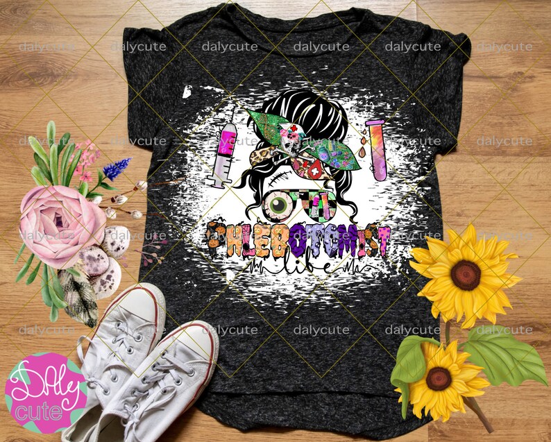 May include: A dark gray heathered t-shirt featuring a graphic of a messy bun hairstyle with a patterned headband, a syringe, a test tube, and glasses. The text PHLEBOTOMIST life is printed below the graphic in a colorful, patterned font.