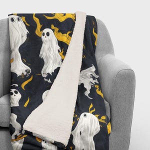 May include: A black and white fleece throw blanket with a pattern of white ghosts and yellow flames. The blanket is folded in half and draped over a gray armchair.