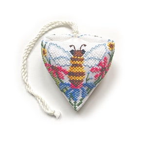 Puede incluir: Una almohada blanca en forma de corazón con un diseño de punto de cruz de una abeja rodeada de flores. La almohada tiene una cuerda blanca atada a la parte superior.