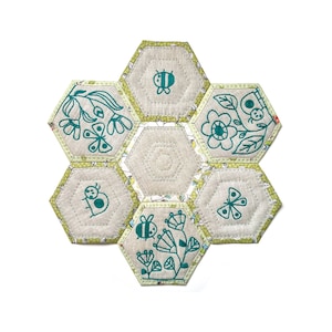 Puede incluir: Una colcha de retazos hexagonal con fondo beige y costuras verdes. Cada hexágono presenta un diseño bordado diferente en verde azulado, incluyendo una abeille, una mariposa, una mariquita y una flor.