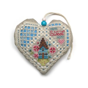 Op de afbeelding: Een hartvormige, beige stoffen ornament met een blauw en wit geborduurd ontwerp. De ornament heeft een klein huis met bloemen en de tekst "Home Sweet Home".
