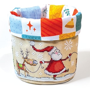 Könnte beinhalten: Ein zylindrischer Aufbewahrungskorb zum Thema Weihnachten. Die Außenseite ist beige mit einem festlichen Weihnachtsmann- und Rentier-Design. Der Rand hat ein farbenfrohes Patchworkmuster mit Sternen, Schneeflocken und geometrischen Formen. Das Innenfutter ist weiß.