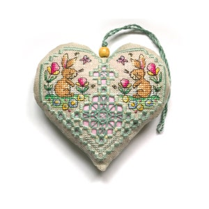 Op de afbeelding: Een hartvormige, beige stoffen ornament met een groen en roze geborduurd ontwerp. Twee bruine konijnen zijn geborduurd op de ornament, omringd door bloemen en vlinders. Een houten kraal en een groene string zijn bevestigd aan de bovenkant van de ornament.