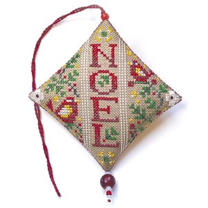 Op de afbeelding: Een ruitvormige, handgestikte kerstversiering met het woord "NOEL" in rode letters. De versiering heeft een beige achtergrond met groene, rode en gele bloemen- en bladerpatronen. Een rode draad en een kralenhangertje maken het ontwerp compleet.