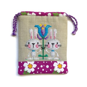 May include: A small, beige drawstring bag with a cross-stitch design of two white bunnies holding a purple flower. The bag has a pink and white polka dot fabric trim.
