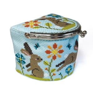 Può includere: Un porta monete in tessuto blu con una chiusura in metallo. Il porta monete è decorato con un ricamo a punto croce di due conigli marroni in un campo di fiori.