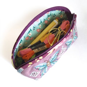 May include: A purple zippered pouch with a floral lining and a butterfly design. The pouch is filled with embroidery floss and a pen.