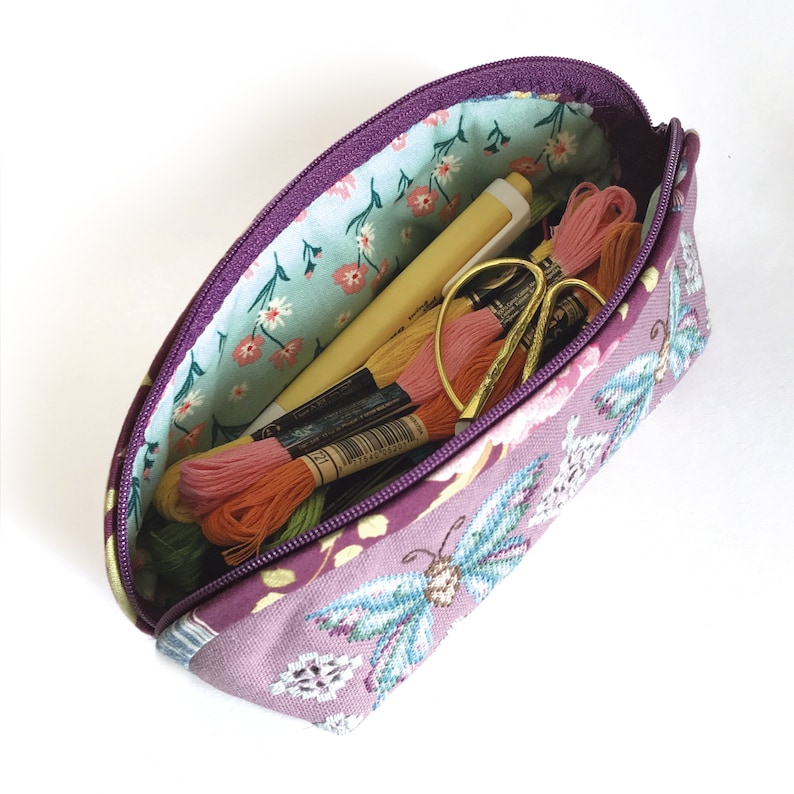 May include: A purple zippered pouch with a floral lining and a butterfly design. The pouch is filled with embroidery floss and a pen.