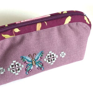 May include: A purple zippered pouch with a floral pattern and a butterfly embroidered on the front. The pouch is made of a soft, textured fabric.