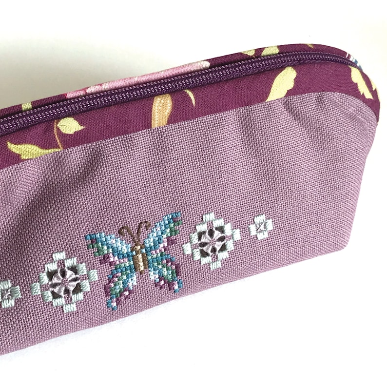 May include: A purple zippered pouch with a floral pattern and a butterfly embroidered on the front. The pouch is made of a soft, textured fabric.