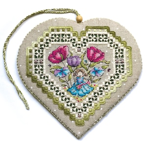 Op de afbeelding: Een hartvormig, kruissteekornament met een bloemenontwerp. Het ornament heeft een centraal beeld van een meisje omringd door roze, paarse en blauwe bloemen. De rand is een gedetailleerd groen en wit patroon en heeft een groen ophangkoord.