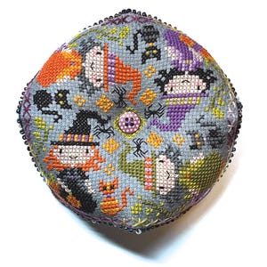 May include: A circular, grey, cross-stitched Halloween pincushion. The design features cartoon witches, cats, bats, and spiders in orange, purple, and black. The edges are trimmed with beads, and a button is in the centre.