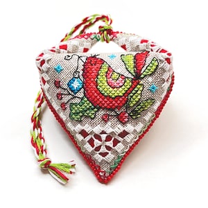 Può includere: Un ornamento fatto a mano a forma di cuore con un design di uccello rosso e verde, circondato da bacche di agrifoglio e un ornamento blu. L'ornamento è realizzato in tessuto di lino e ha un cordino intrecciato rosso e bianco.