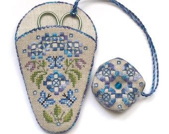 Puede incluir: Un protector de agujas y un porta tijeras a juego, ambos con un diseño floral azul y blanco. El porta tijeras tiene un lazo para colgar y un par de tijeras en su interior.
