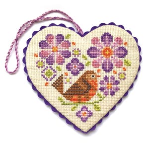 May include: Heart-shaped cross-stitch decoration featuring a brown bird perched on a branch, surrounded by purple and pink flowers. The edges are trimmed with a scalloped purple border, and a matching cord is attached for hanging.