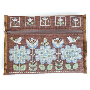 Può includere: Una pochette con cerniera marrone con un motivo floreale ricamato sul davanti. I fiori sono blu, bianchi e verdi. Ci sono due uccelli sulla pochette, uno su ogni lato dei fiori.